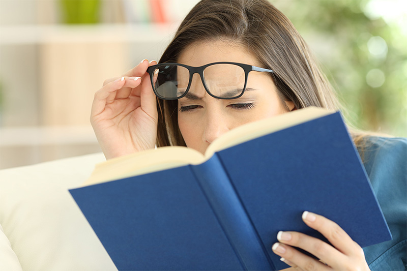 A woman is holding her glasses while reading a book and squinting, as if having difficulty seeing the text clearly.